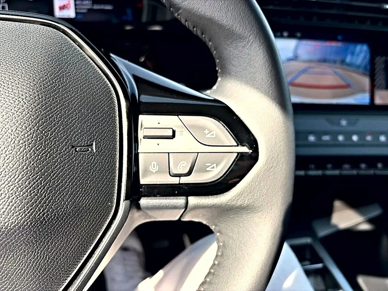 Gros plan sur le volant cuir noir de la Peugeot 308 2025 avec commandes audio et écran de caméra de recul en arrière-plan.