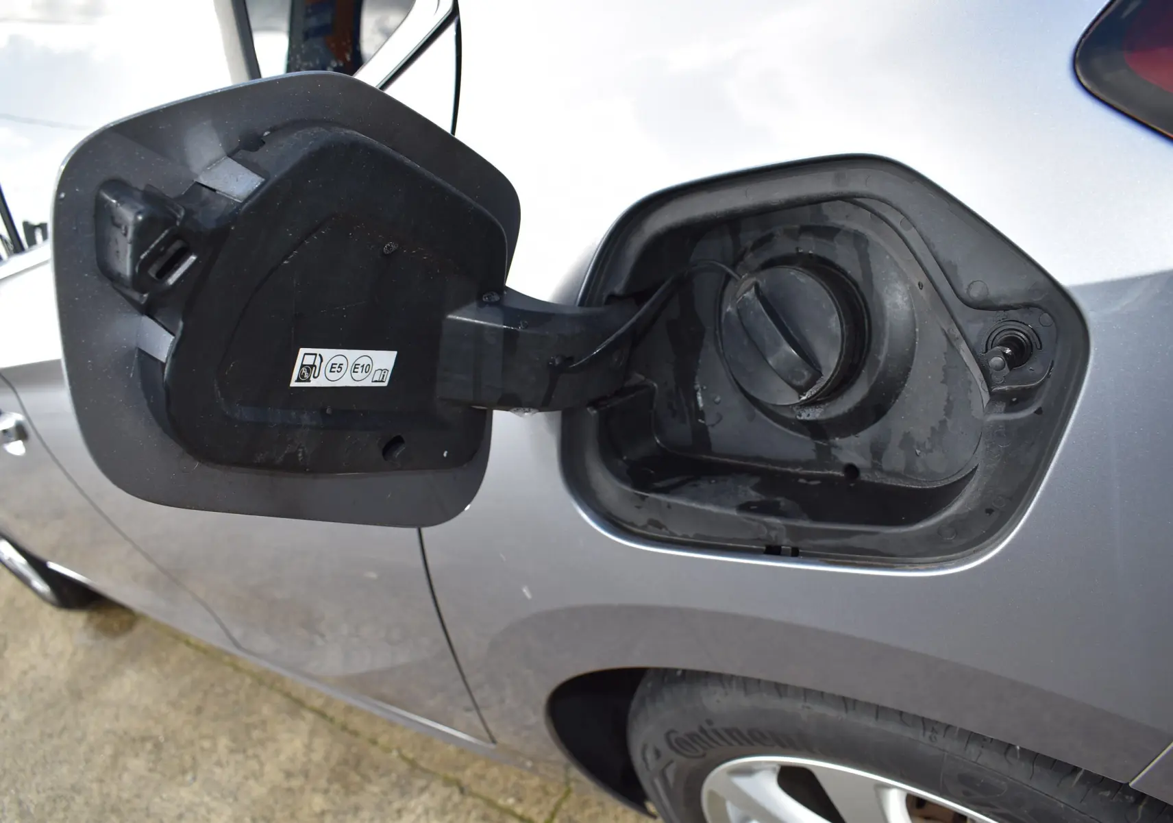 Ouverture de la trappe à carburant côté gauche d'une Opel Corsa gris clair essence 2021, avec bouchon visible.