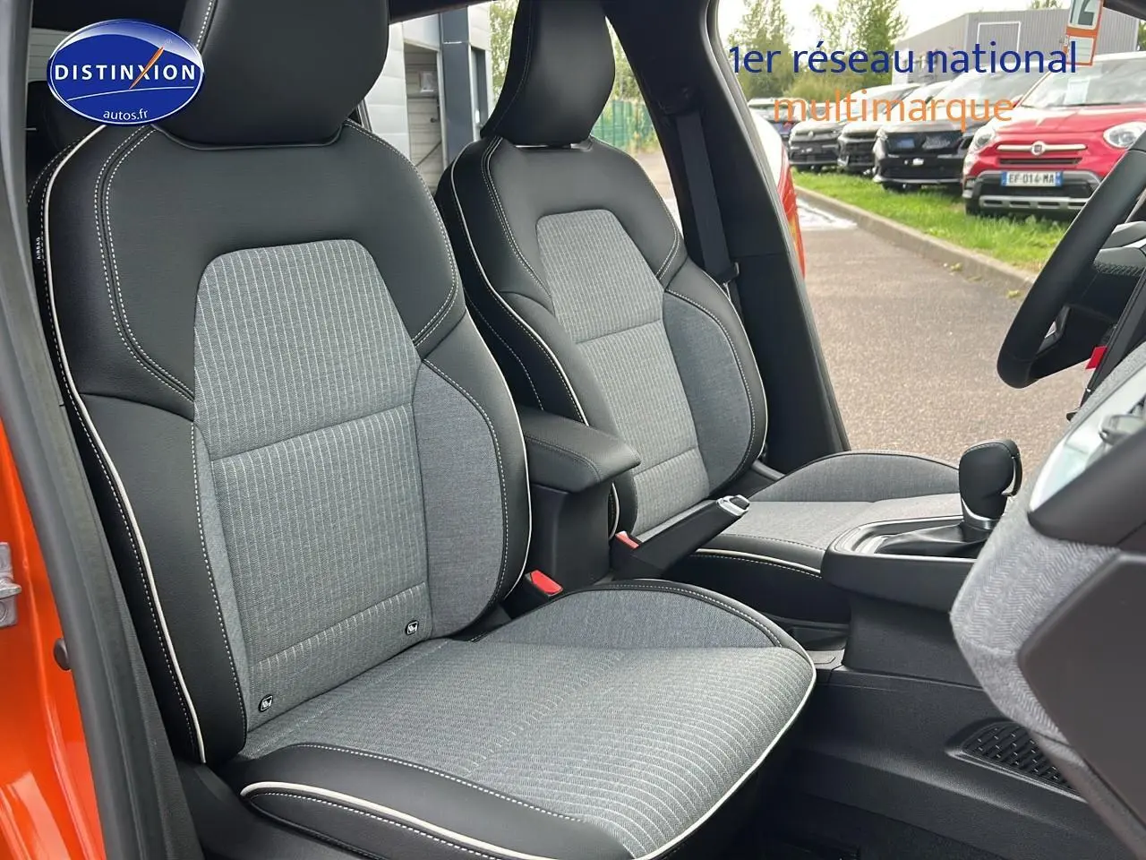 Vue intérieure côté conducteur de la Renault Clio V orange Valencia, sièges sport gris avec surpiqûres blanches et accoudoir central.