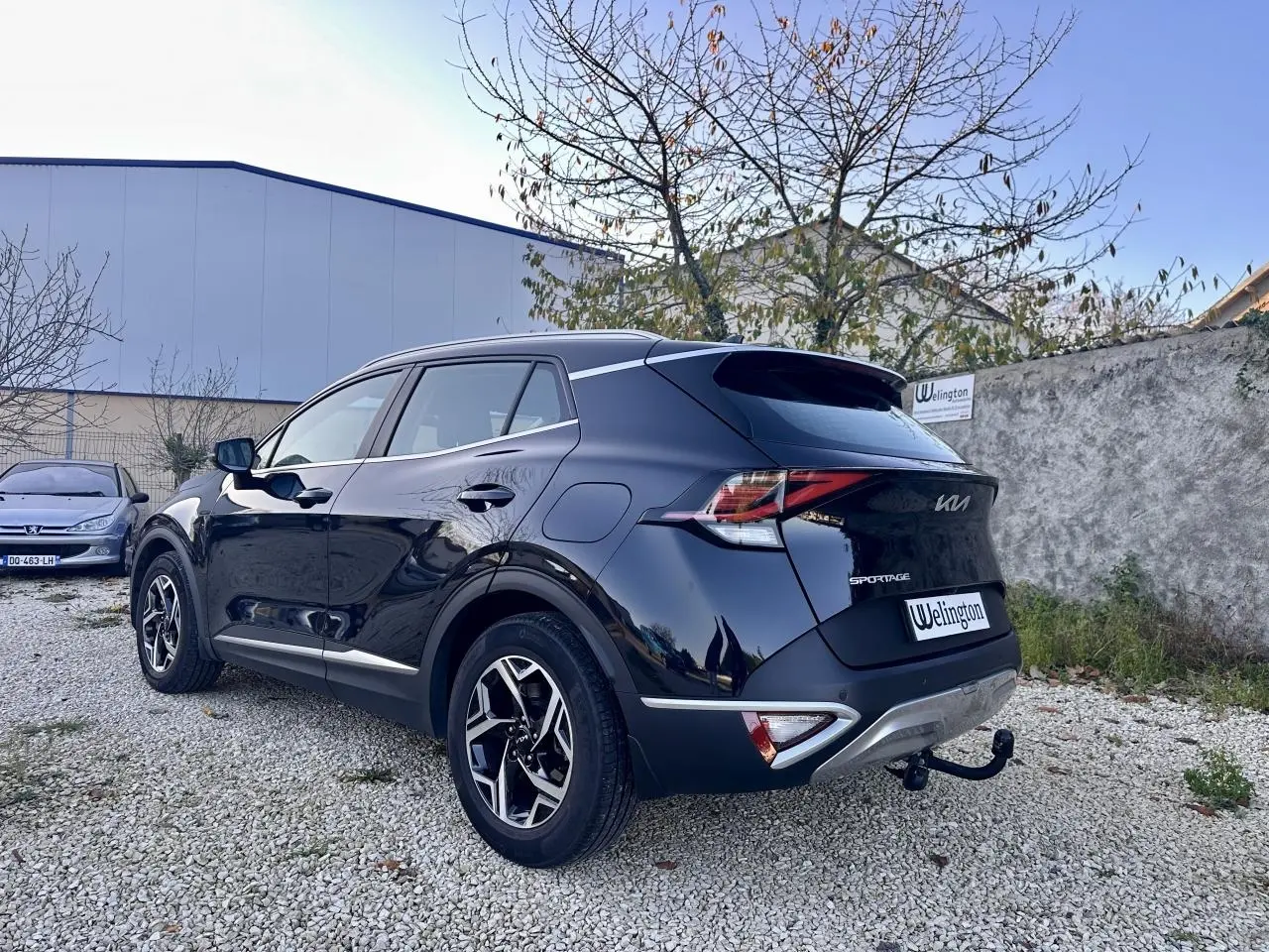 Vue 3/4 arrière droit du Kia Sportage noir 2023 avec attelage et jantes alliage distinctives sur un sol gravillonné.