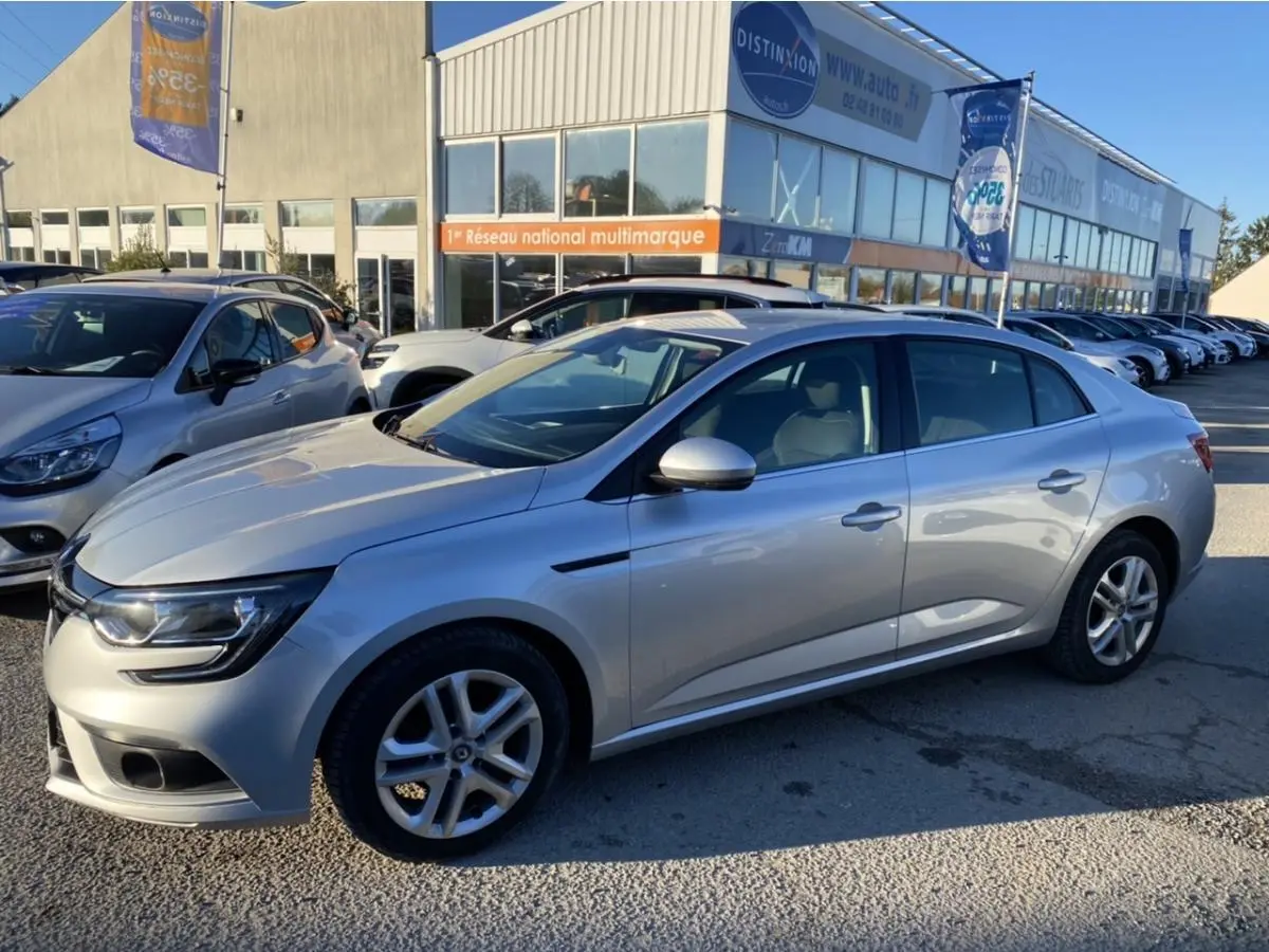 Renault Mégane Sedan gris platine vue de profil côté droit, stationnée devant un concessionnaire multimarque.