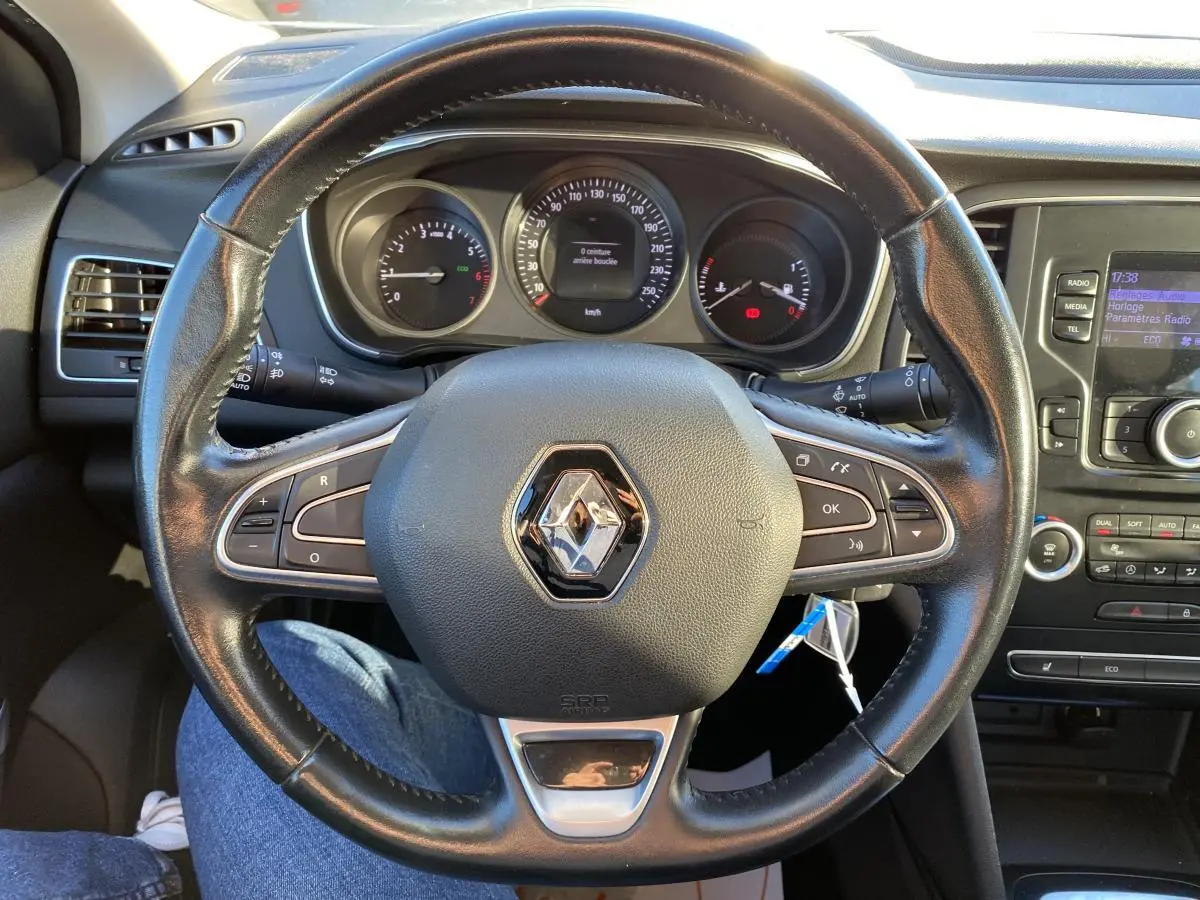 Vue rapprochée du volant en cuir noir avec logo Renault, tableau de bord et console centrale d'une Mégane gris platine 2017.