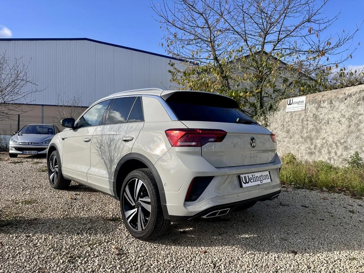 Volkswagen T-Roc gris clair vue 3/4 arrière droit, avec jantes alliage et vitres teintées sur parking extérieur.