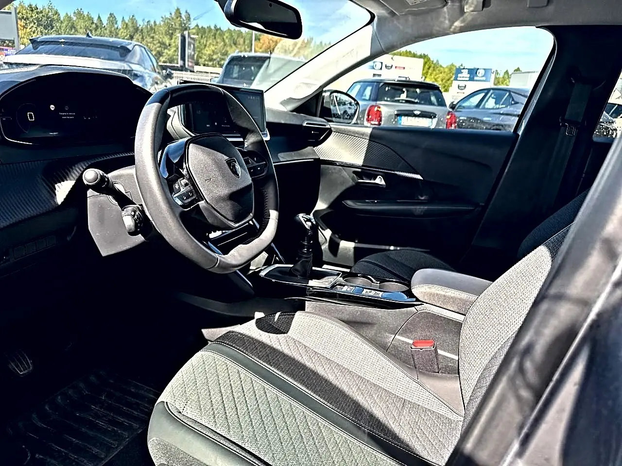 Vue intérieure côté conducteur de la Peugeot 208 1.2 100 Allure 2025 avec sellerie tissu gris et volant compact multifonction.
