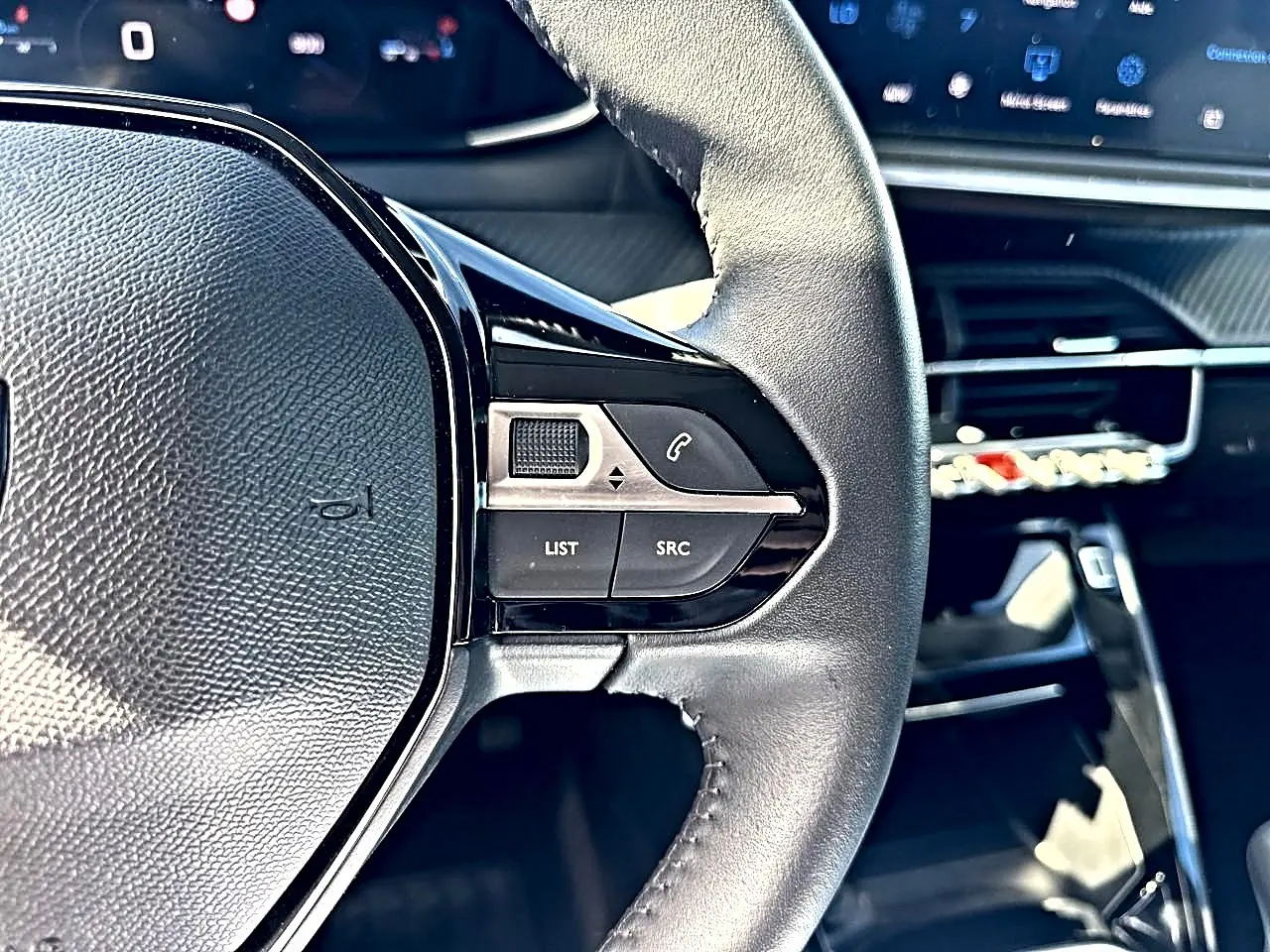 Gros plan sur le volant cuir noir de la Peugeot 208 1.2 100 Allure, avec commandes audio et tableau de bord numérique.