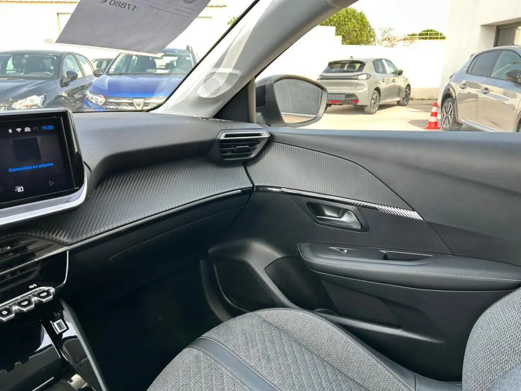 Vue intérieure côté passager de la Peugeot 208 Hybrid 110 e-DCS6 Allure 2024 avec tableau de bord gris et sièges tissu.