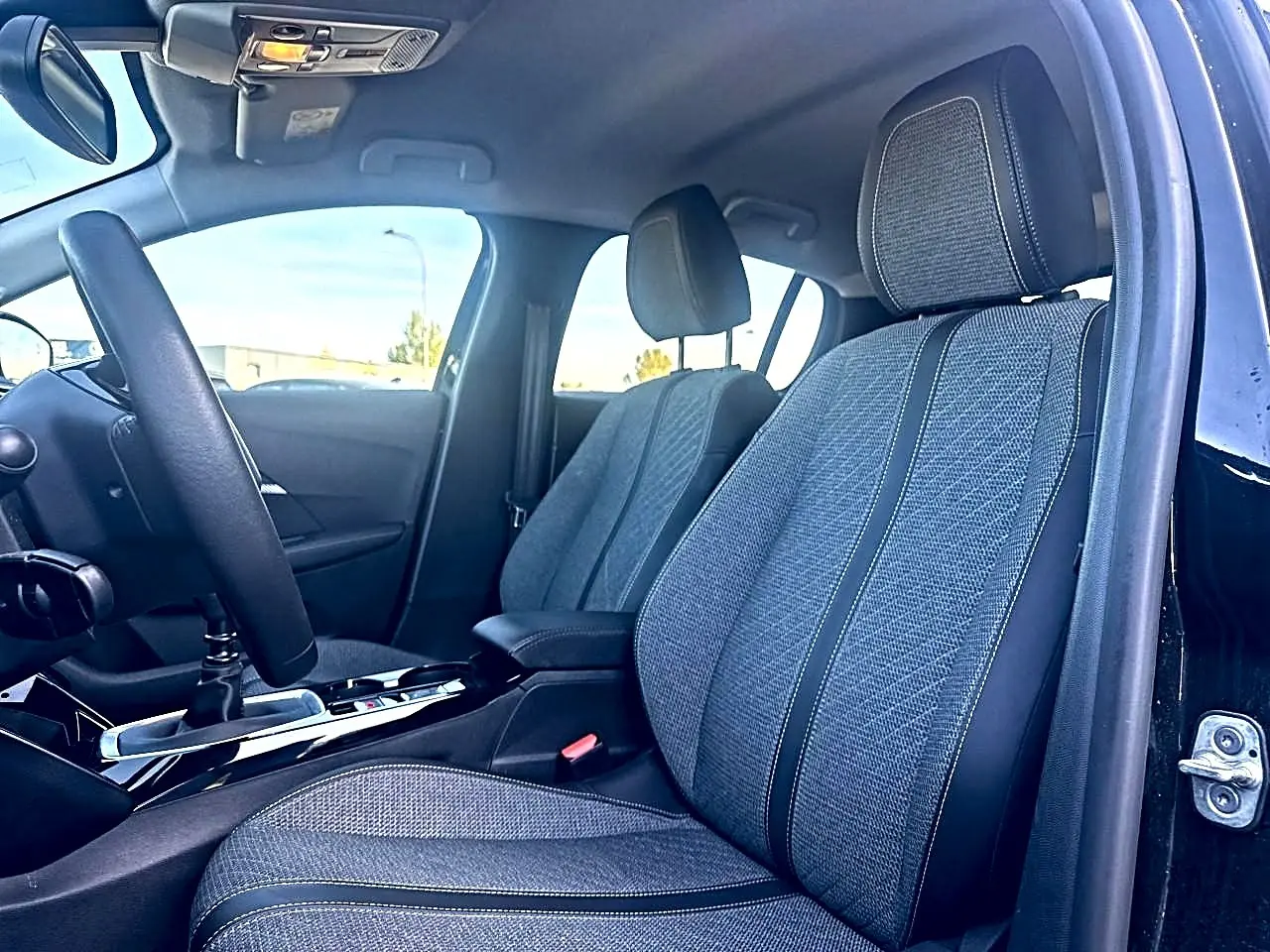 Vue intérieure côté conducteur de la Peugeot 208 noire 2025, mettant en valeur les sièges en tissu gris et le volant noir.