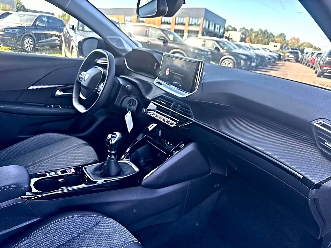 Vue intérieure côté conducteur de la Peugeot 208 noire, avec tableau de bord moderne et boîte manuelle visible.