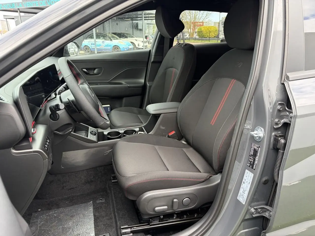 Vue intérieure côté conducteur du Hyundai Kona II 1.6 Hybrid N Line gris Ecotronic avec sièges noirs à surpiqûres rouges.