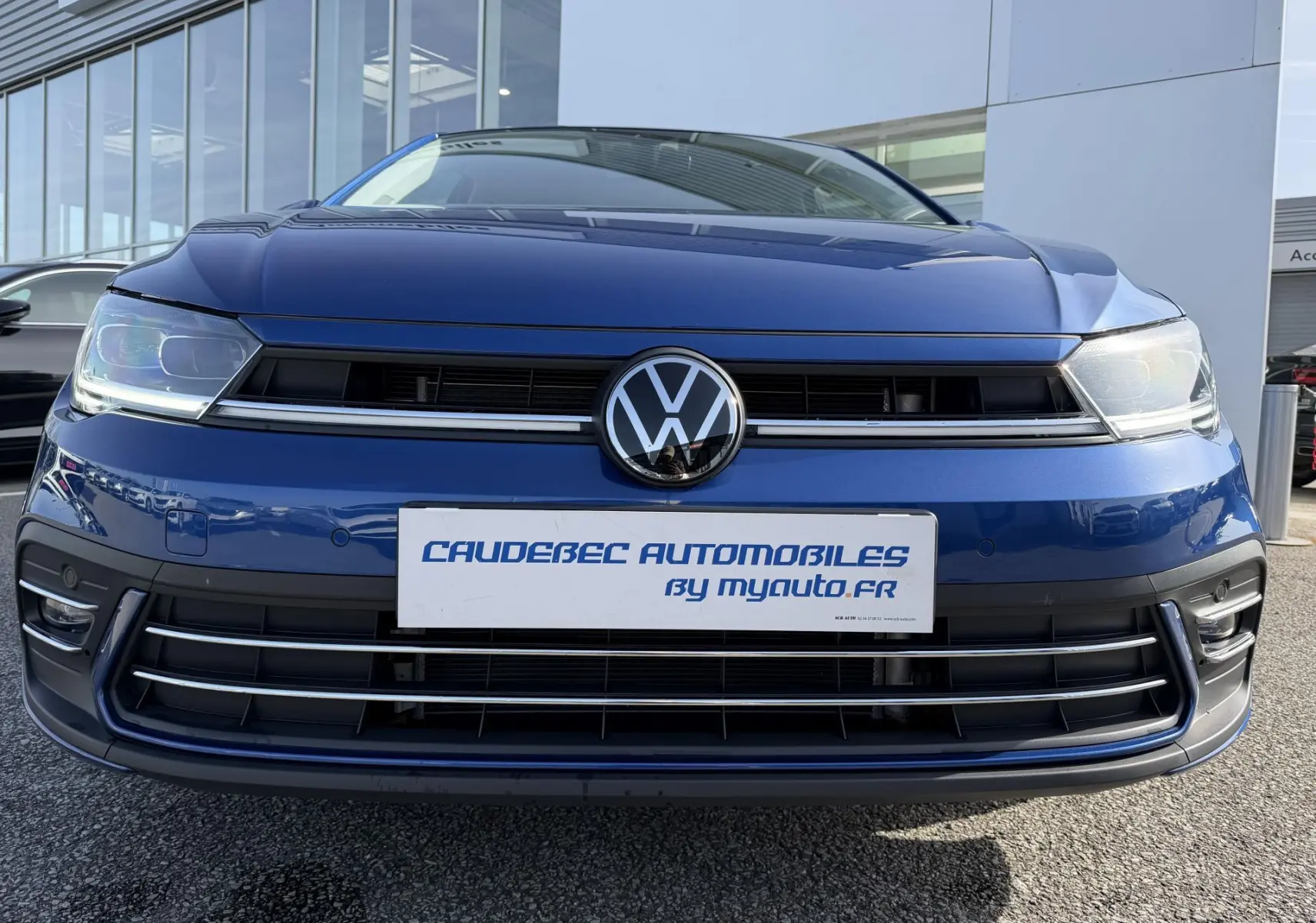 Vue frontale rapprochée d'une Volkswagen Polo 2025 bleu récif métallisé avec logo VW centré et phares LED allumés.