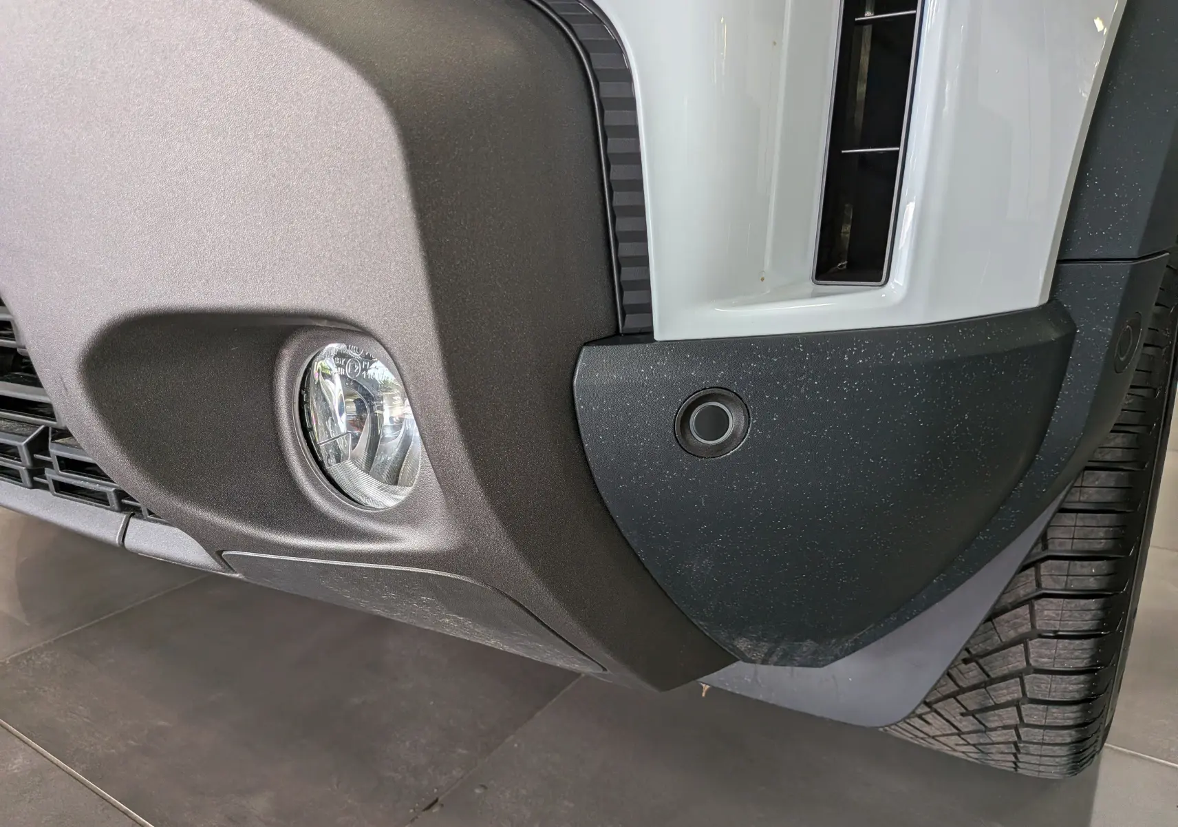 Gros plan sur le pare-chocs avant droit blanc glacier du Dacia Duster Hybrid-G avec antibrouillard et capteur de stationnement.