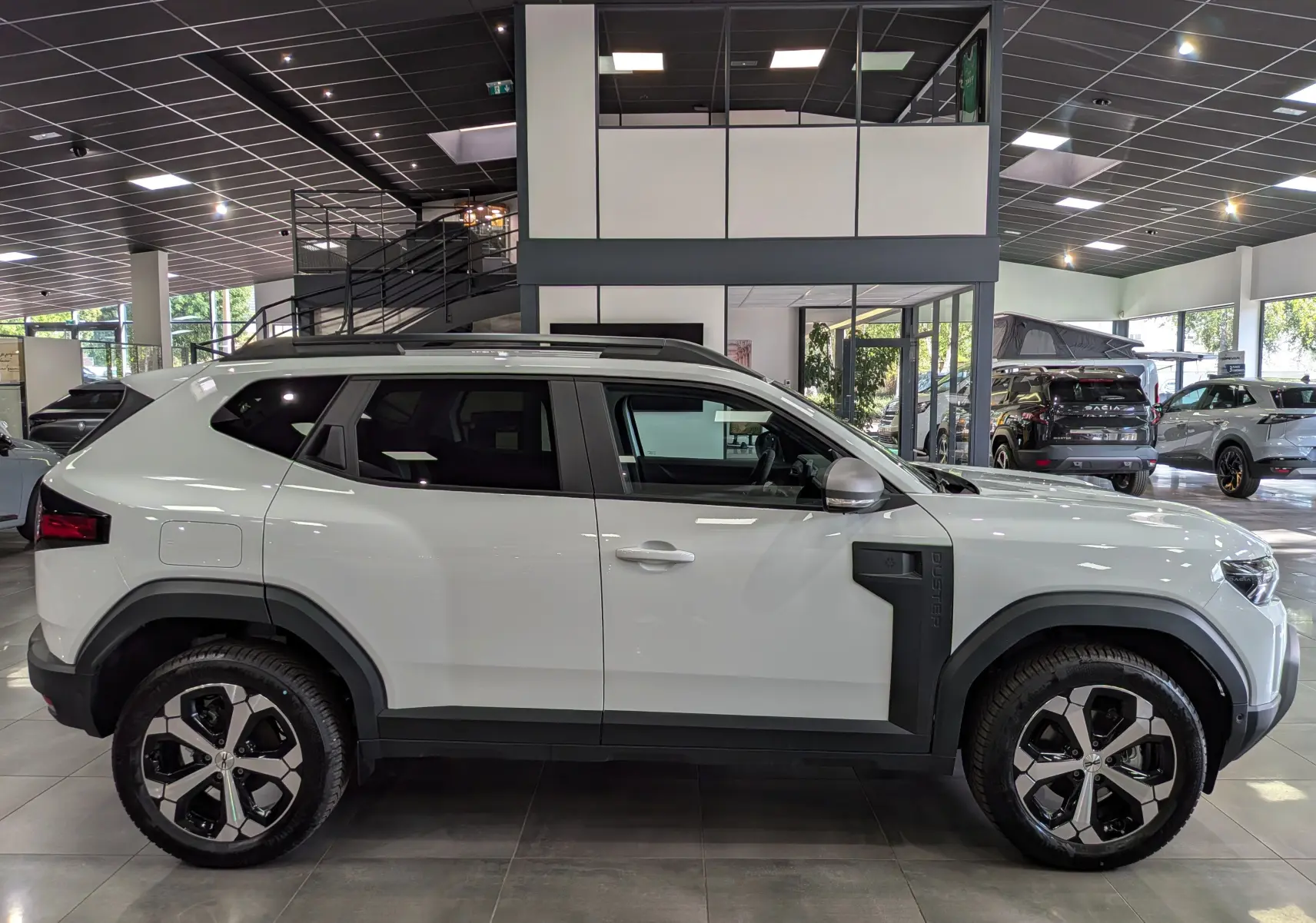 Profil droit du Dacia Duster Hybrid-G blanc glacier 2026 en showroom, avec jantes bicolores et protections latérales noires.