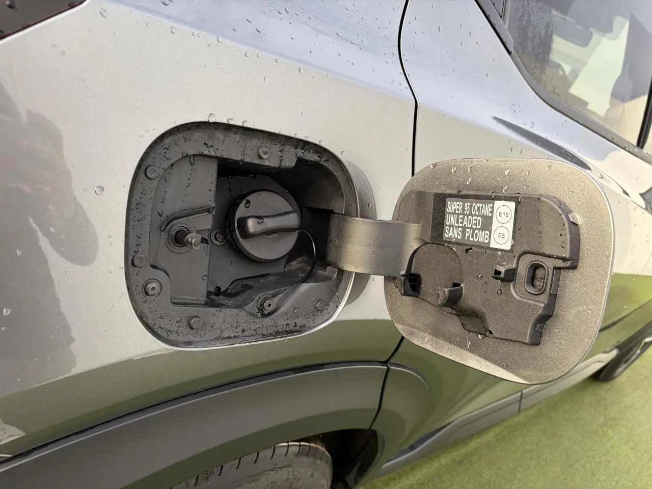 Gros plan sur la trappe à carburant ouverte côté gauche d’un Nissan Juke II noir métal avec pluie visible.