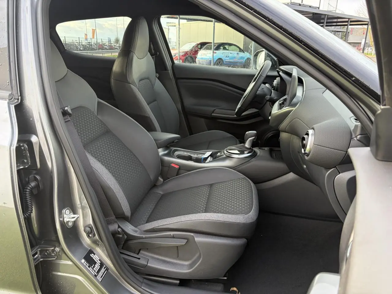 Vue intérieure côté conducteur du Nissan Juke II 1.6 Hybrid noir métal, avec sièges tissu et console centrale moderne.