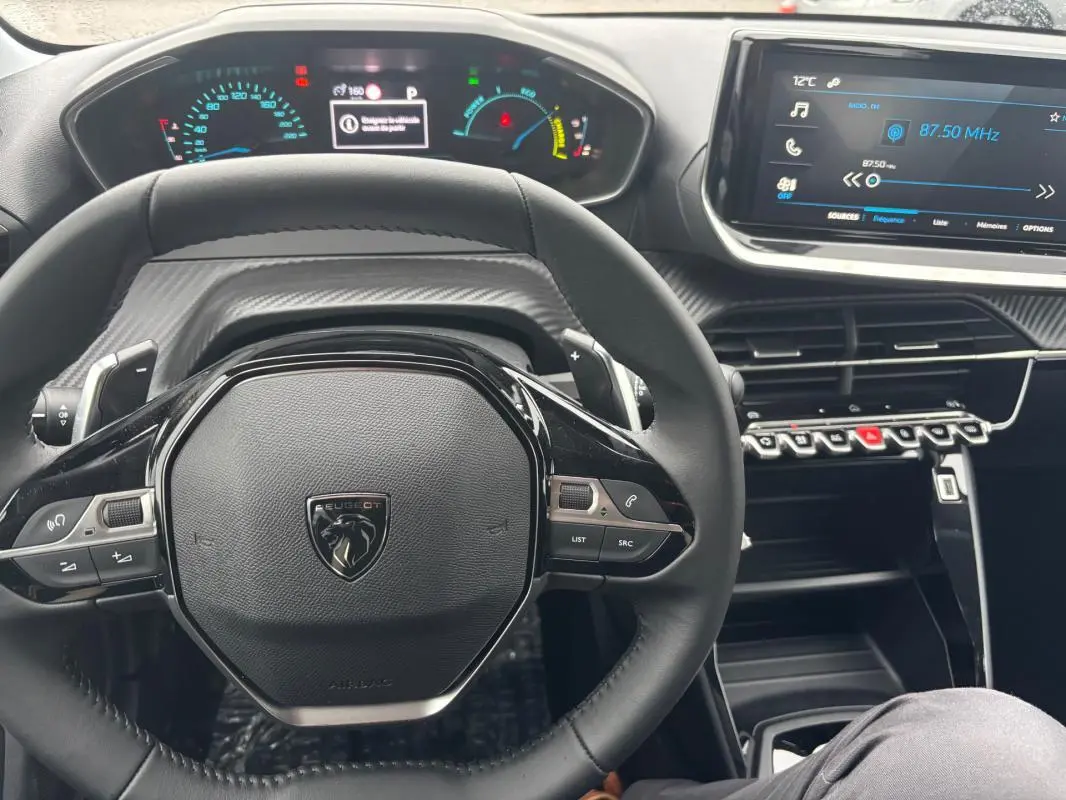 Vue rapprochée du volant noir de la Peugeot 208 Hybrid 2025 avec cockpit numérique et écran tactile allumé.