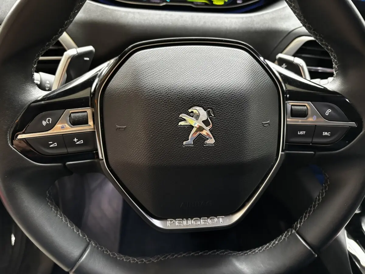 Gros plan sur le volant cuir noir du Peugeot 3008 1.6 HYBRID 225 Allure Pack avec commandes intégrées et logo lion chromé.