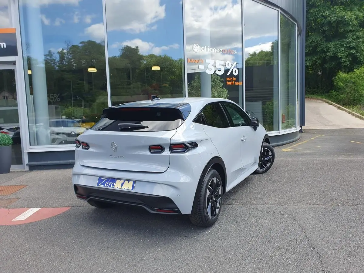 Vue 3/4 arrière droite d'une Renault Clio VI gris Rafale avec jantes alliage noir devant un bâtiment vitré.
