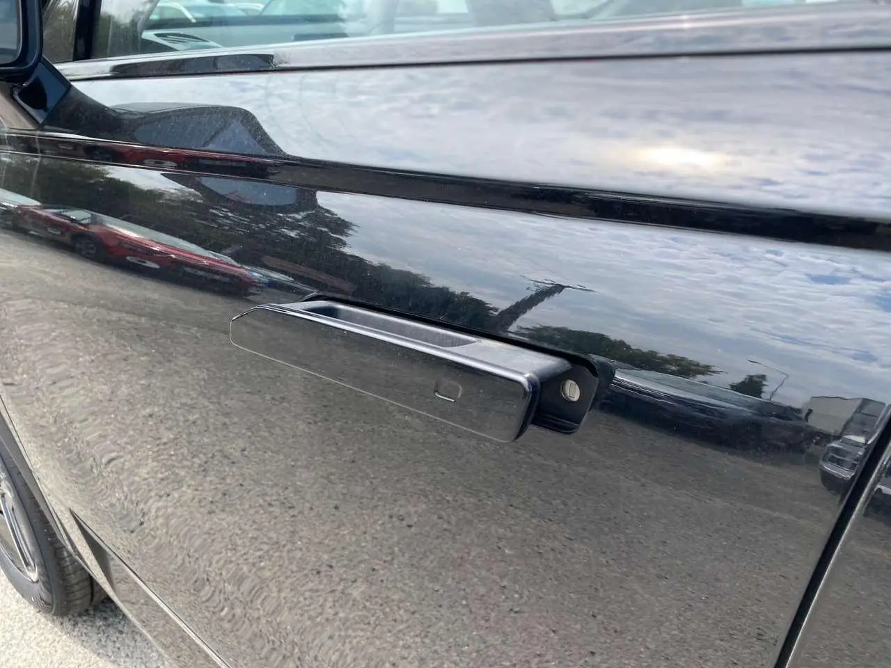 Poignée de porte rétractable côté gauche du JAECOO J7 2026 en noir Carbon Cristal, reflet du ciel et environnement urbain.