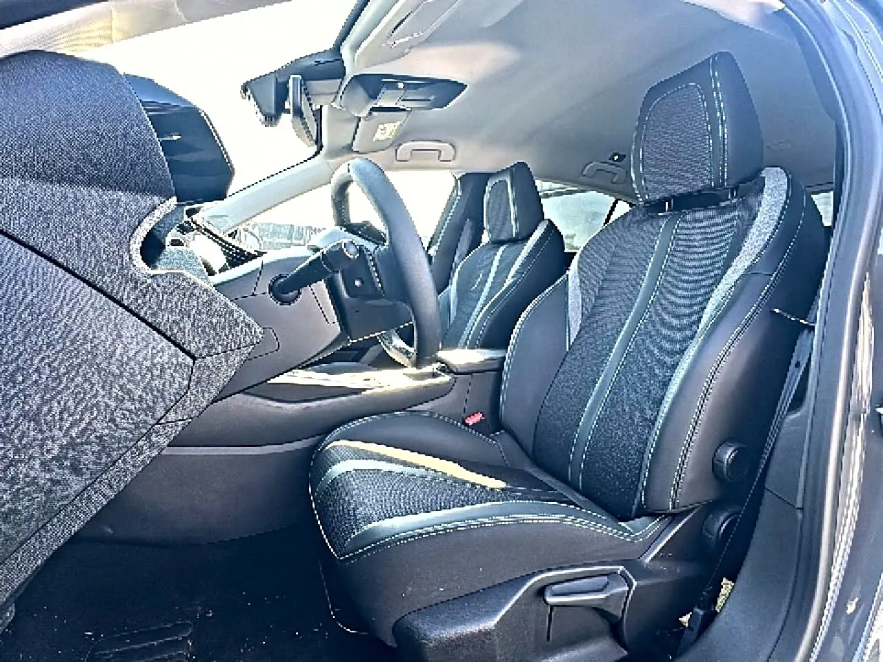 Vue intérieure côté conducteur de la Peugeot 308 2025, sièges tissu noir avec surpiqûres contrastantes et volant multifonction.
