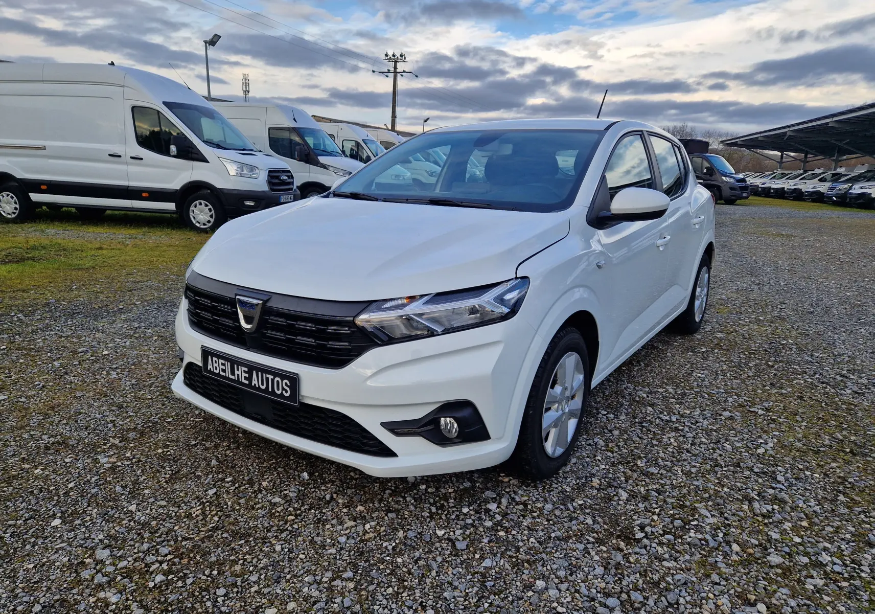 Dacia Sandero blanc vue 3/4 avant droit sur parking gravillonné avec plusieurs fourgons blancs en arrière-plan.
