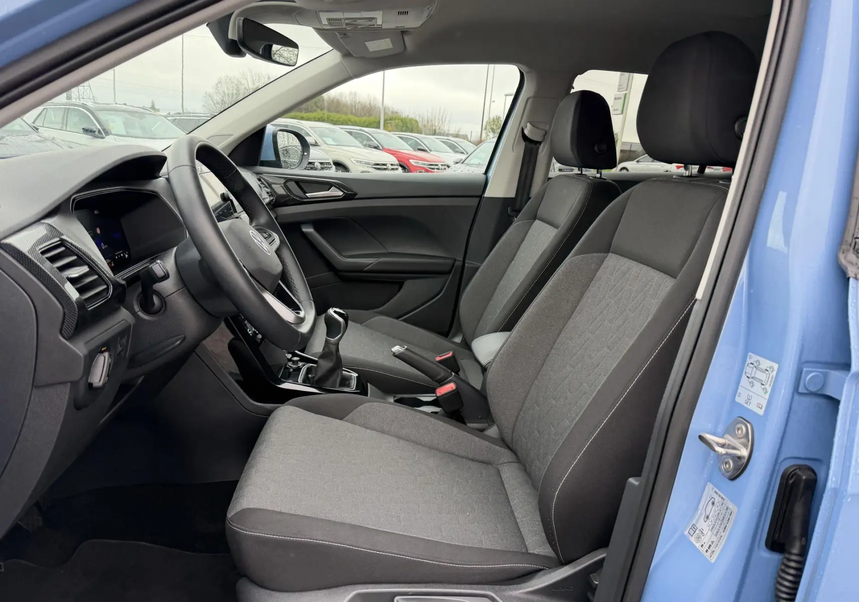Vue intérieure côté gauche du Volkswagen T-Cross bleu 2025, montrant sièges tissu gris et volant multifonction cuir.