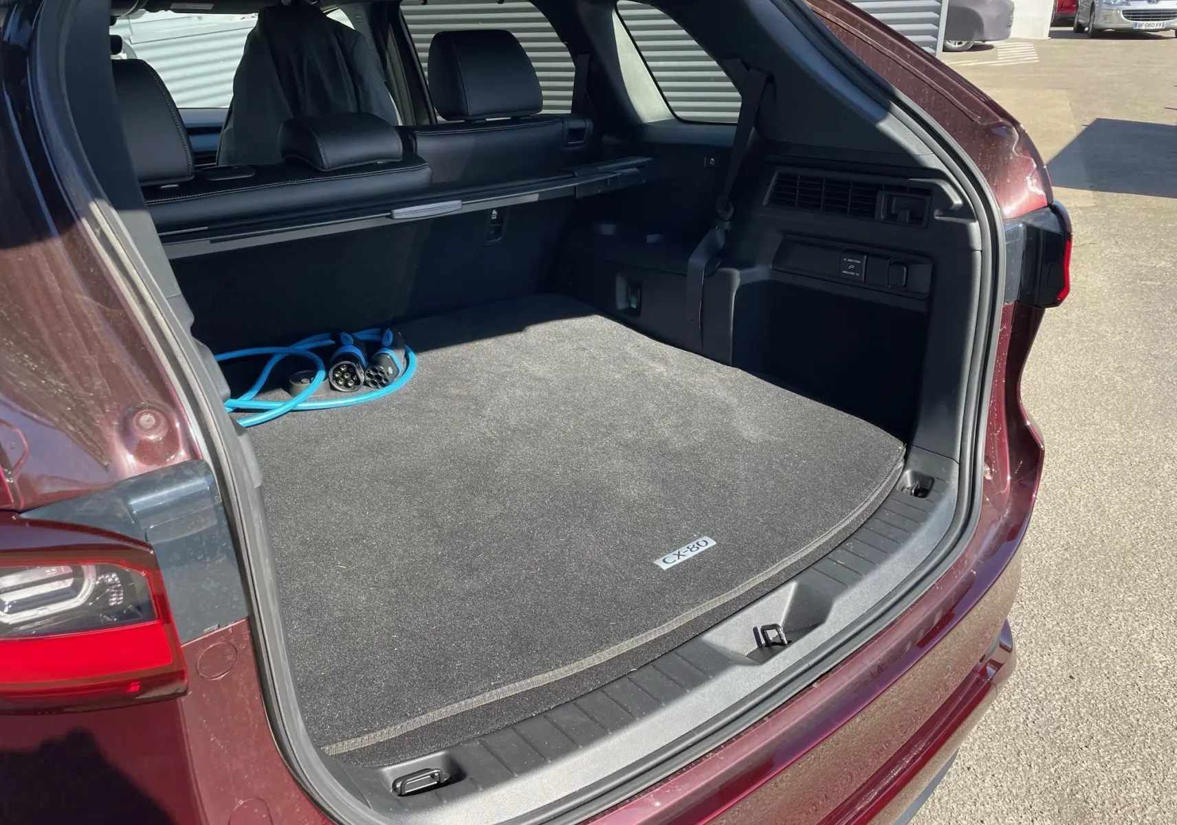 Coffre ouvert du Mazda CX-80 Artisan Red 2024, vue arrière droite avec câble de recharge hybride visible.