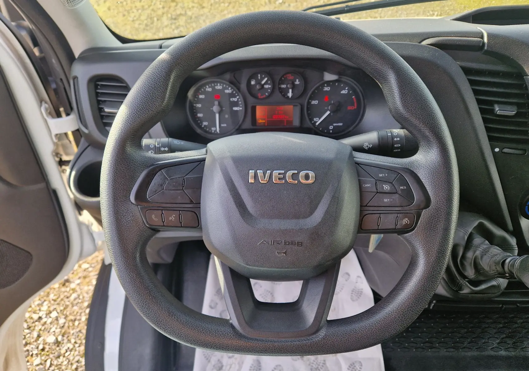 Vue intérieure centrée sur le volant noir d’un Iveco Daily Fourgon blanc, avec tableau de bord et commandes visibles.