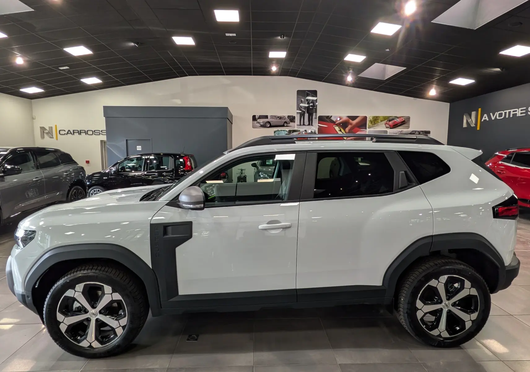 Vue de profil côté gauche d’un Dacia Duster Hybrid-G blanc Glacier avec jantes bi-ton et protections noires contrastantes.