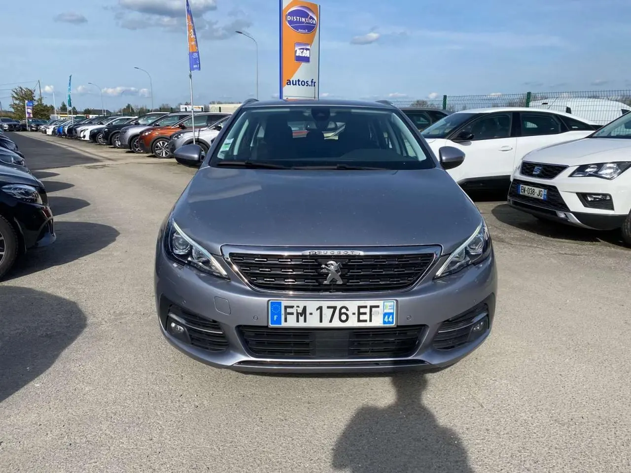 Vue frontale d'une Peugeot 308 SW gris Artense métallisé stationnée sur un parking extérieur avec plusieurs autres véhicules.