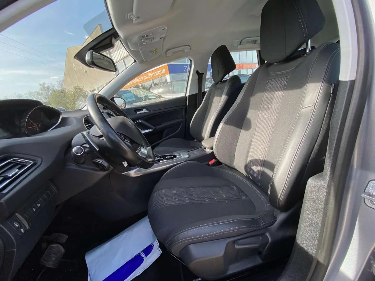 Vue intérieure côté conducteur de la Peugeot 308 SW gris Artense, montrant sièges noirs et tableau de bord moderne.