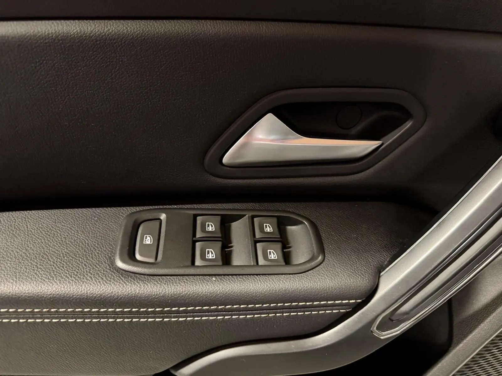 Poignée de porte intérieure noire avec surpiqûres blanches et commandes de vitres électriques côté conducteur d'un Dacia Duster gris.