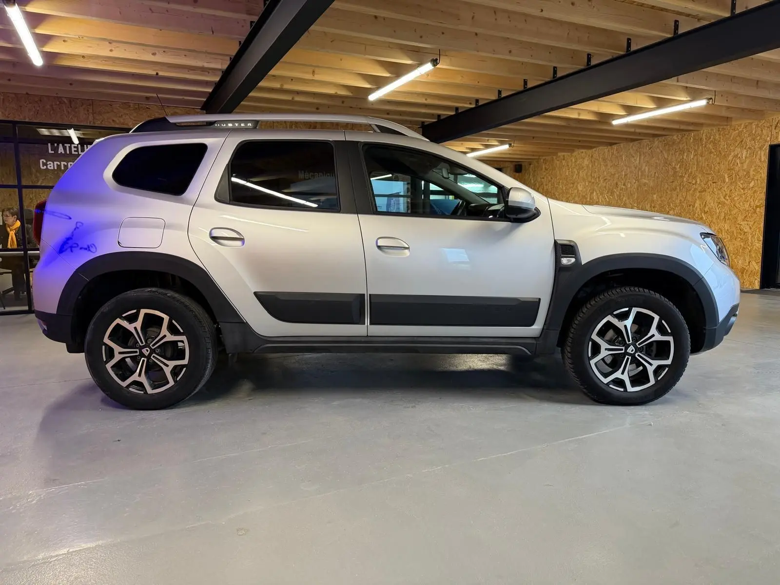 Profil côté gauche d'un Dacia Duster gris 2019 avec jantes alliage 17'' Maldives et barres de toit visibles.