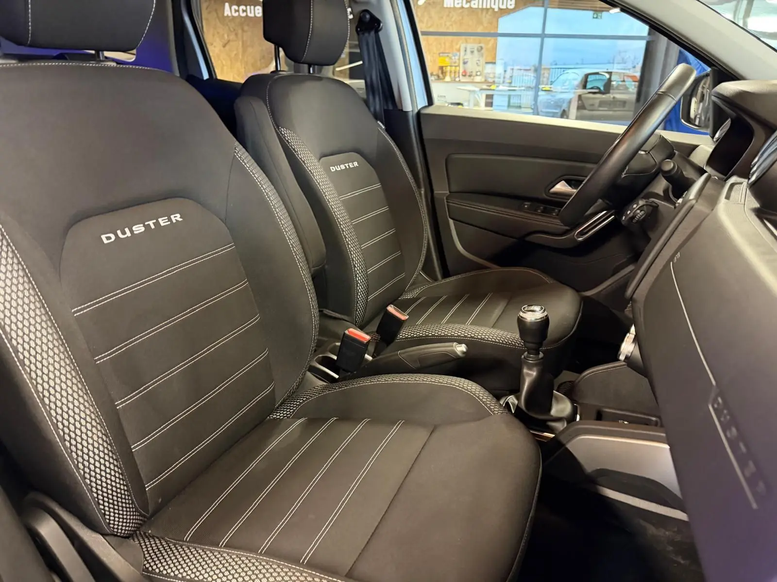 Intérieur avant du Dacia Duster 2019, sièges noirs "Duster" avec surpiqûres, volant et levier de vitesse visibles côté droit.