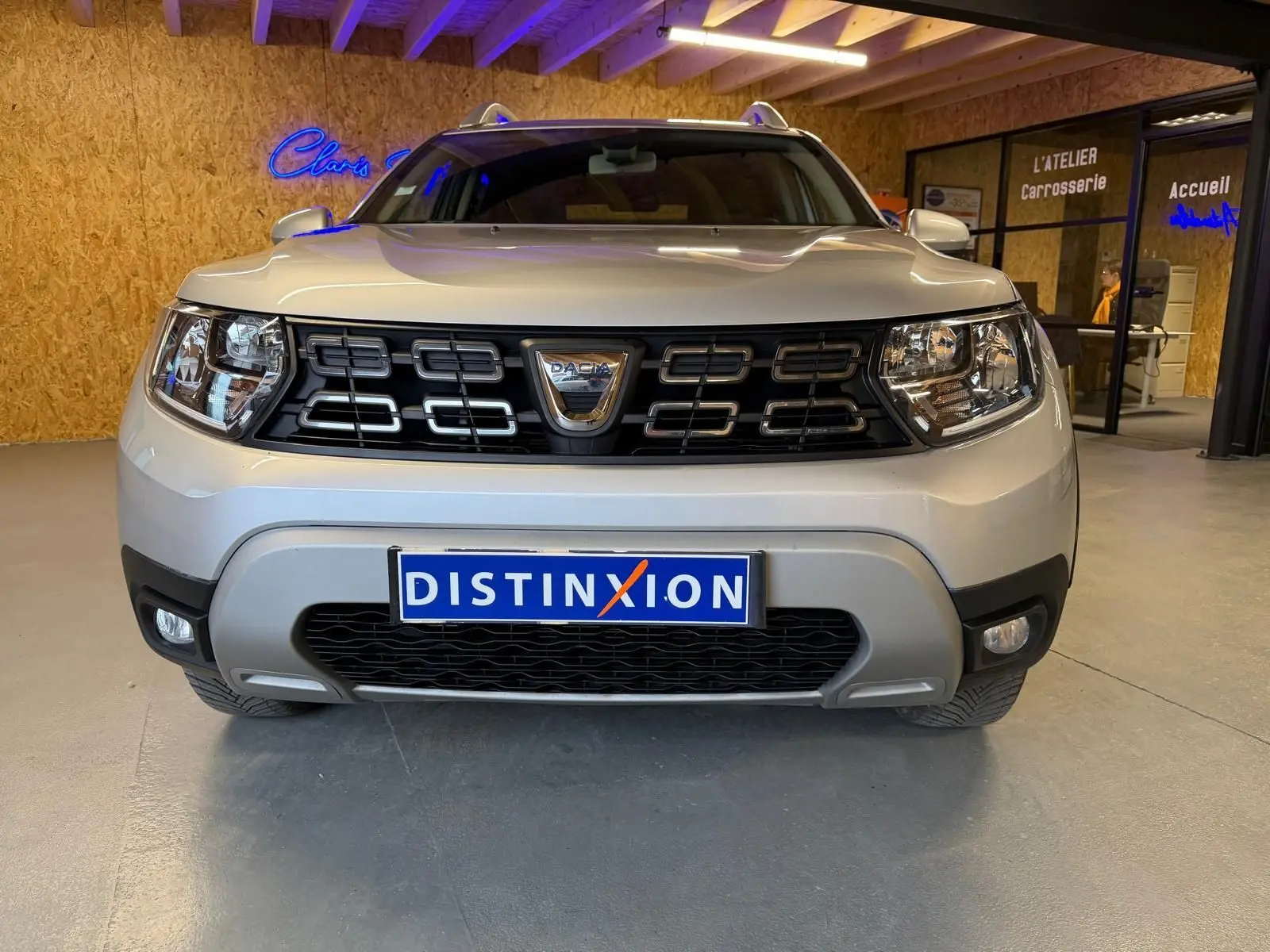Vue frontale d'un Dacia Duster gris 2019 avec calandre noire et logo chromé, dans un atelier lumineux.
