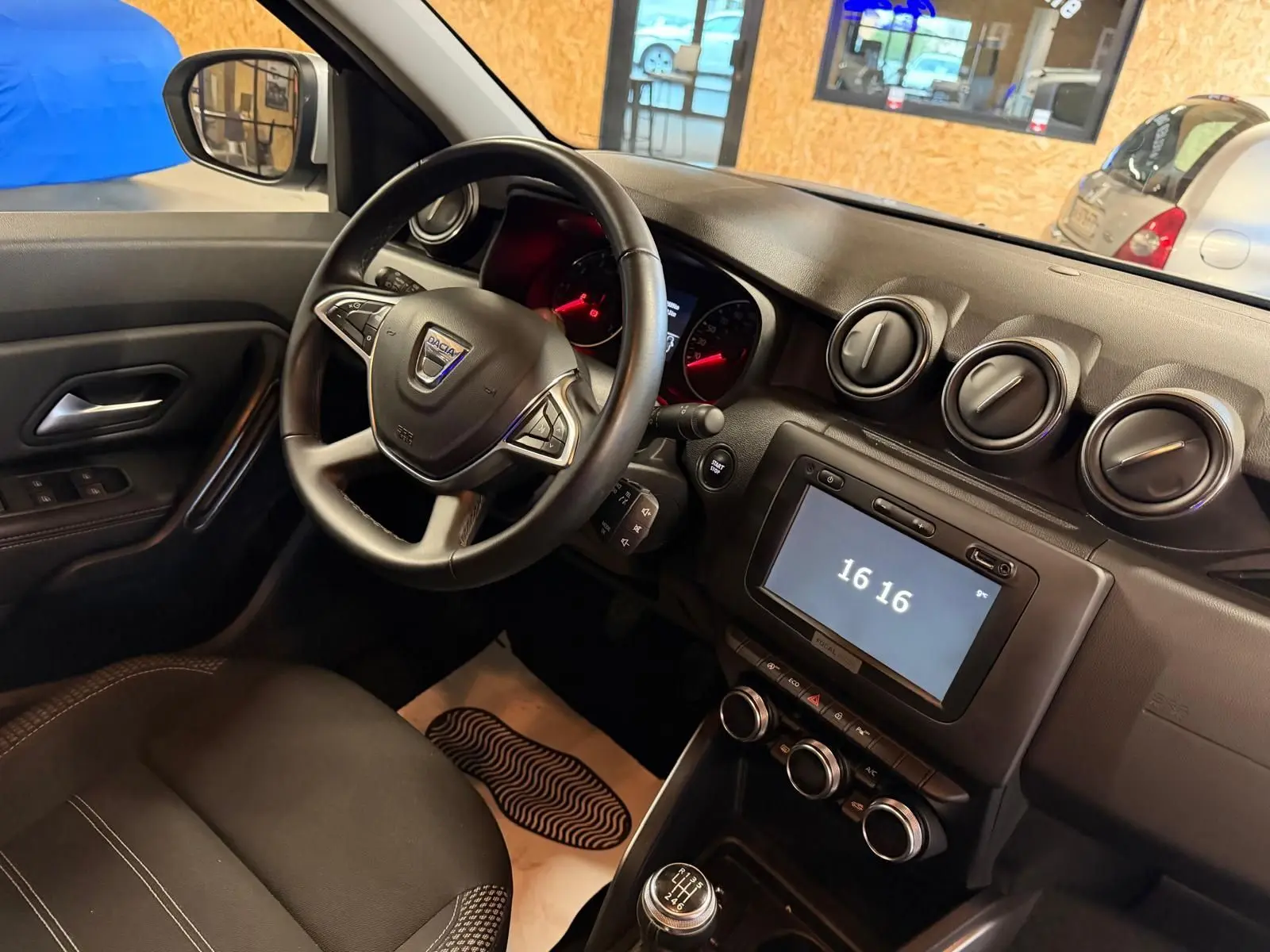 Vue intérieure côté conducteur du Dacia Duster gris 2019, volant multifonction et tableau de bord avec écran tactile allumé à 16h16.