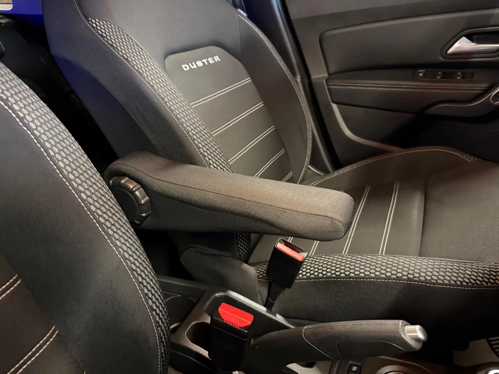Gros plan sur le siège conducteur noir avec accoudoir et logo DUSTER, côté gauche intérieur d'un Dacia Duster gris.