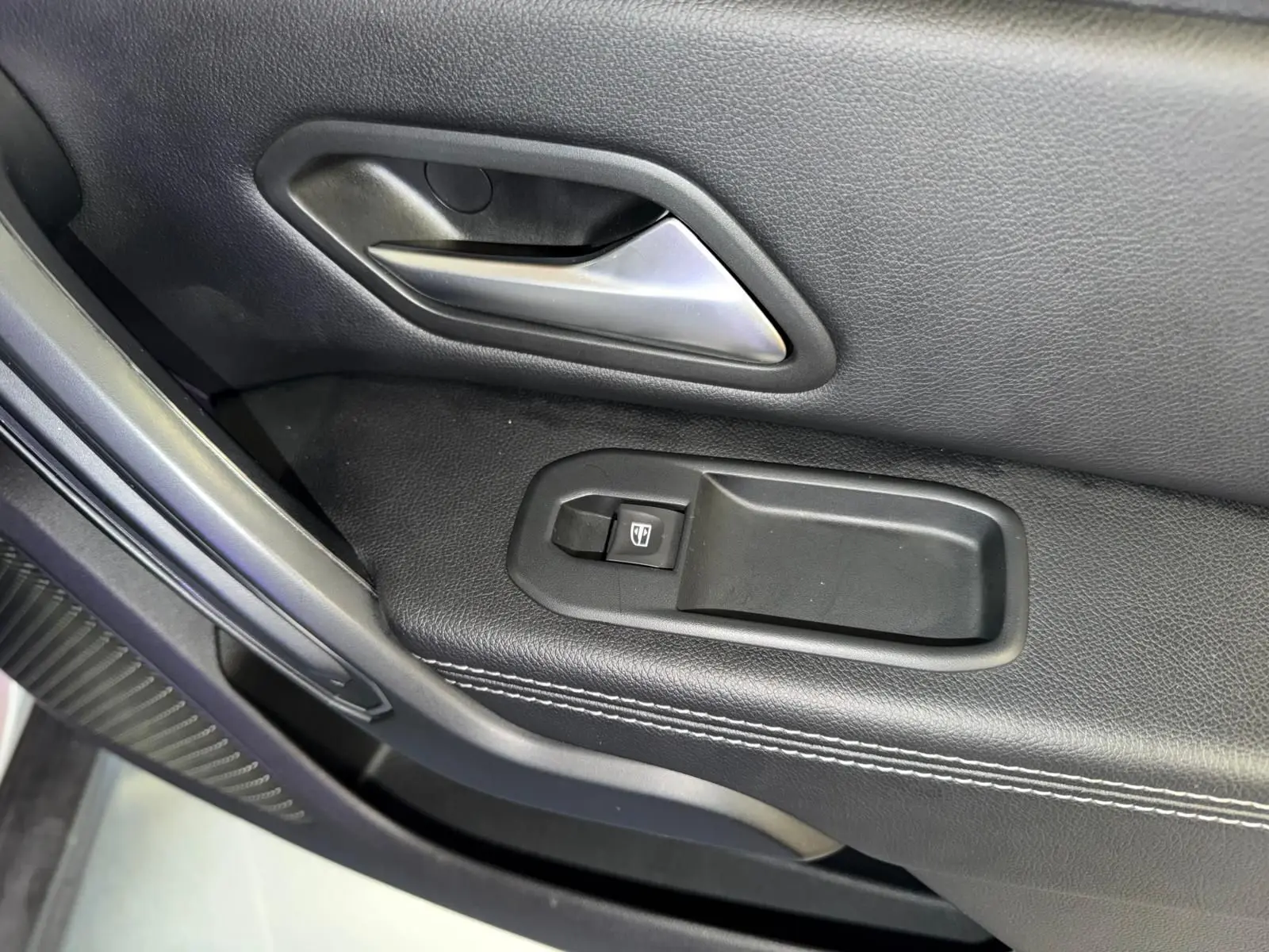 Gros plan sur la commande de lève-vitre électrique et la poignée intérieure chromée côté conducteur du Dacia Duster gris.