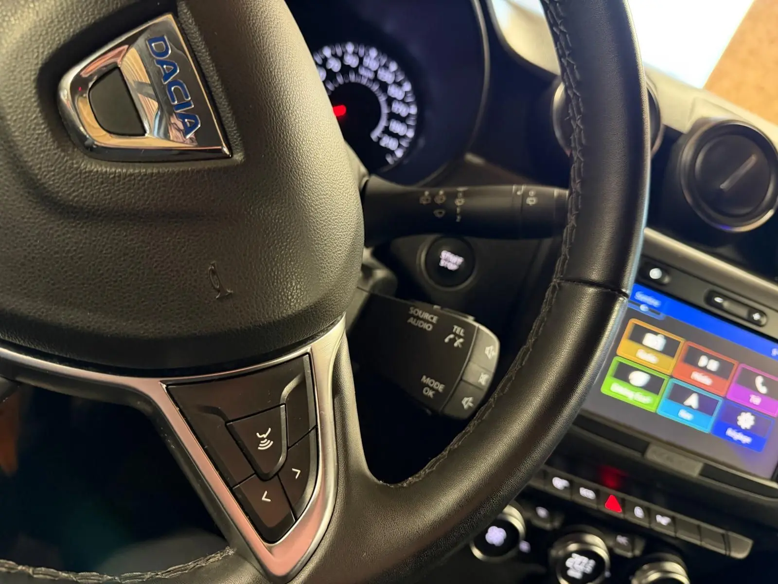 Gros plan sur le volant en cuir noir du Dacia Duster gris, avec commandes audio et tableau de bord lumineux en arrière-plan.