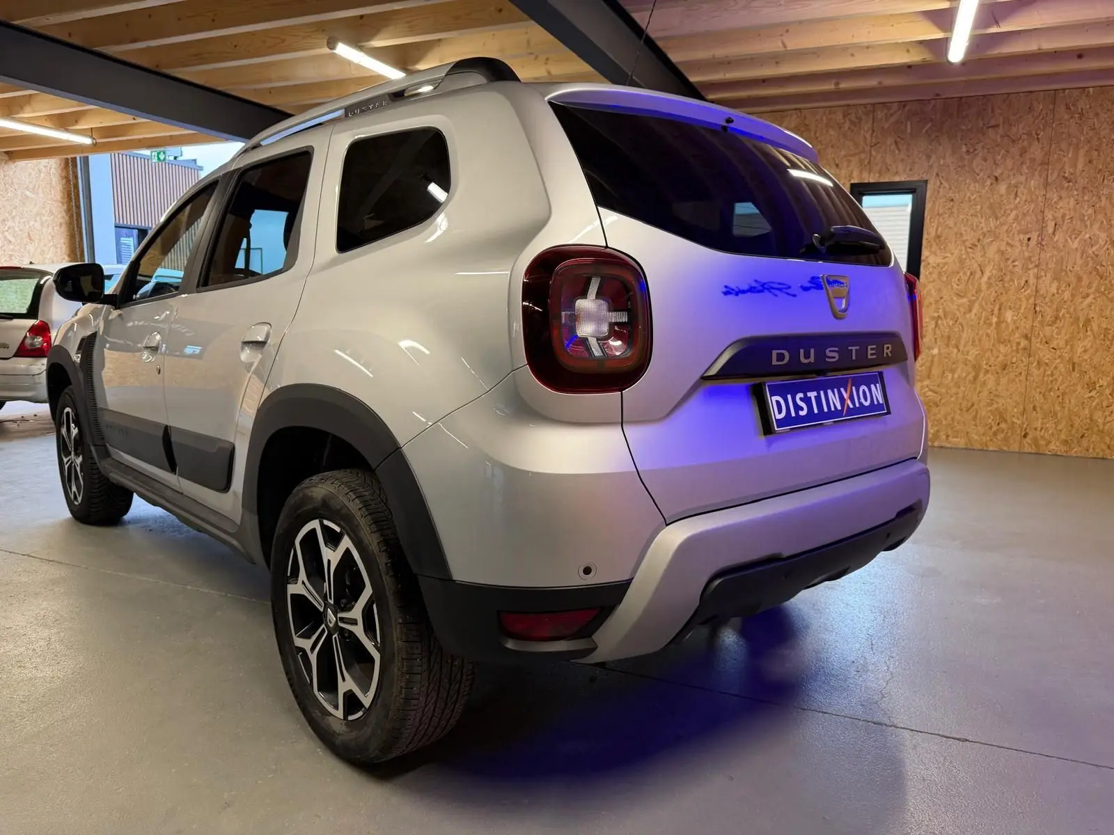 Vue 3/4 arrière droit d'un Dacia Duster gris 2019 avec jantes alliage et barres de toit en intérieur d'atelier.