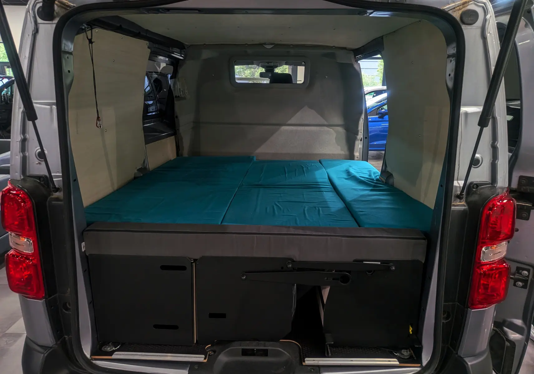Vue arrière ouverte du Peugeot Expert Fourgon gris acier 2023, aménagé avec un lit bleu et habillage bois intérieur.