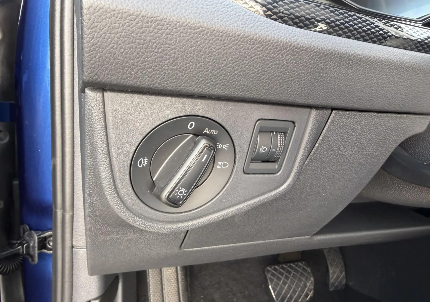 Vue intérieure côté gauche du tableau de bord bleu du Volkswagen Taigo 2025, avec commande des phares et réglage de l'éclairage.