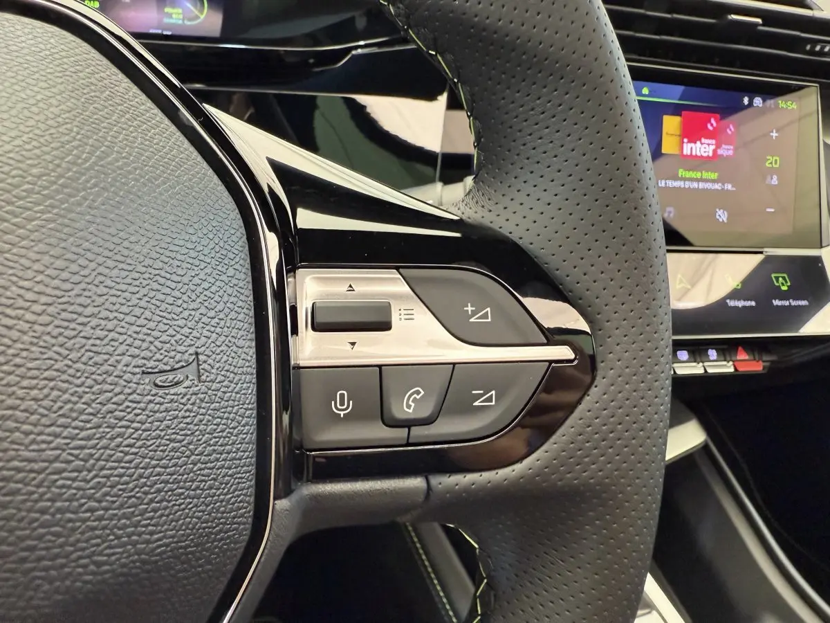 Gros plan sur le volant cuir perforé de la Peugeot 308 Hybrid gris foncé, avec commandes vocales et écran tactile en arrière-plan.