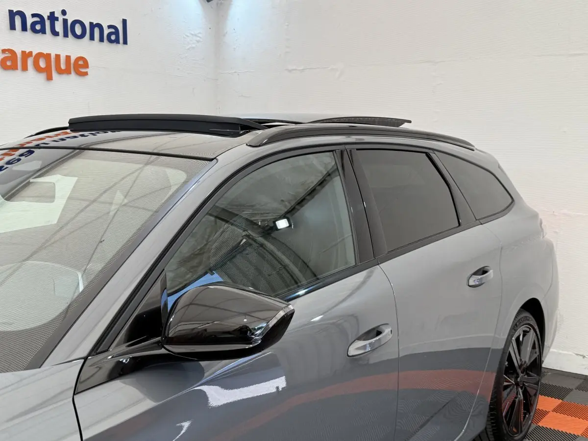 Vue côté gauche avant d'une Peugeot 308 Hybrid 136 e-DCS6 GT gris foncé avec rétroviseur noir brillant et barres de toit.