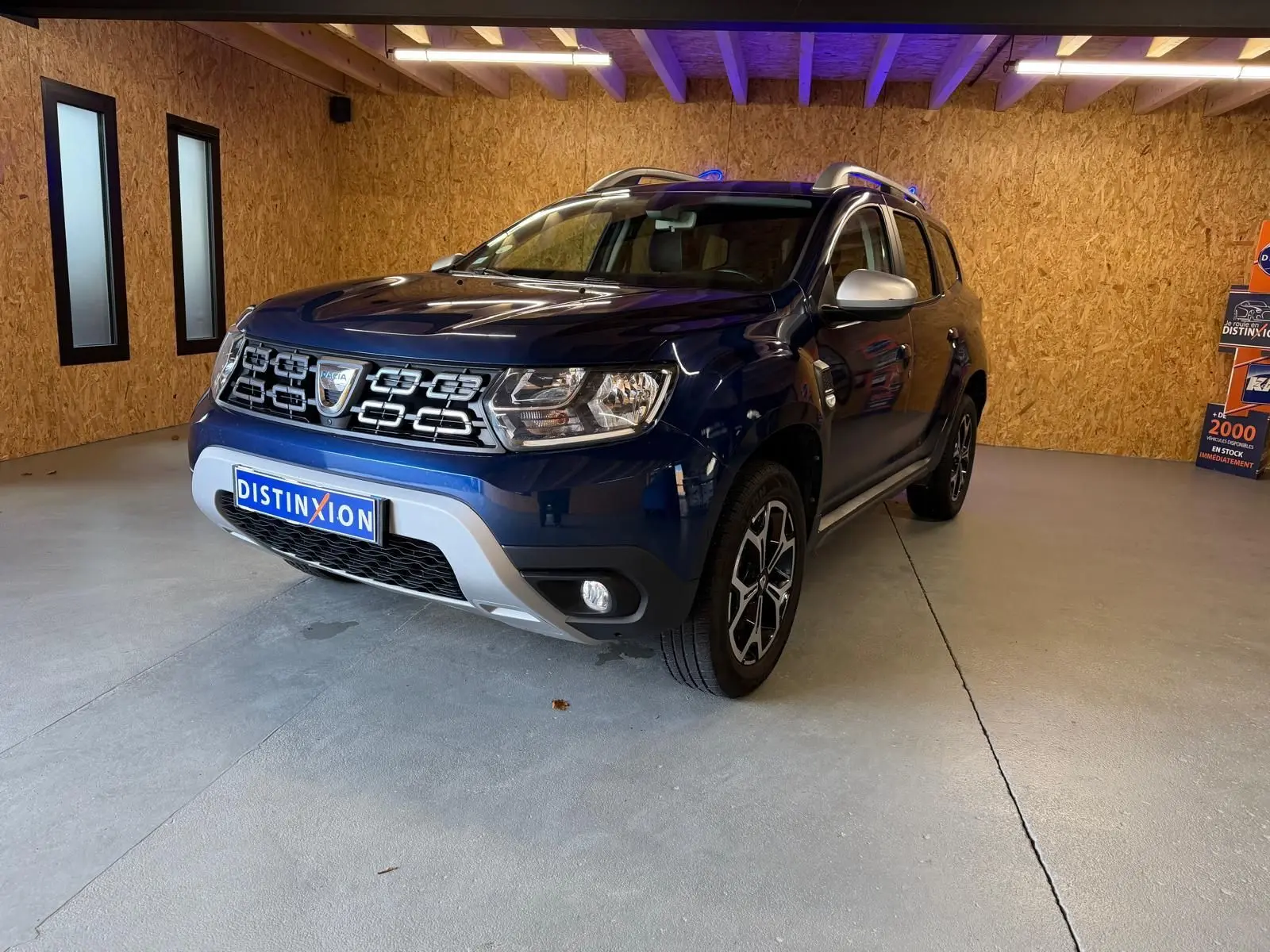 Vue 3/4 avant d'un Dacia Duster bleu avec calandre chromée et jantes alliage 17 pouces dans un garage.