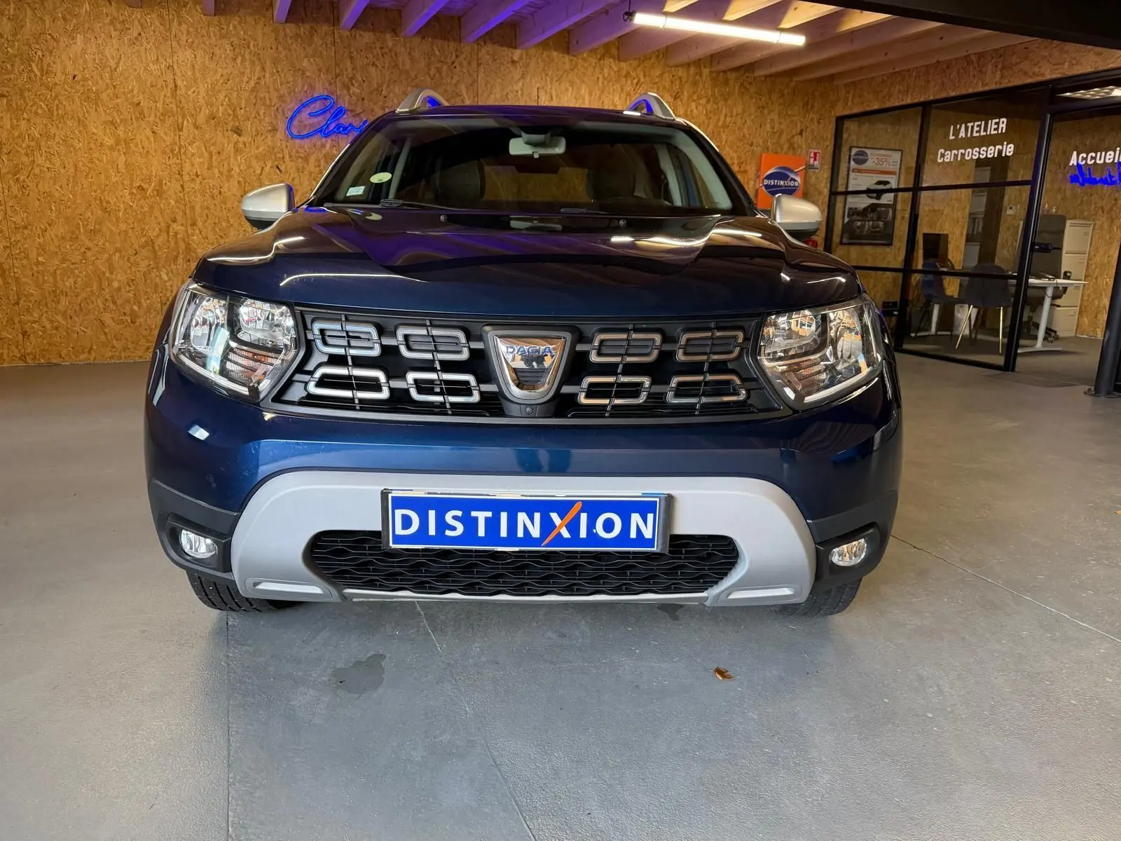 Vue frontale d'un Dacia Duster bleu avec calandre noire et éléments chromés, stationné en intérieur showroom.