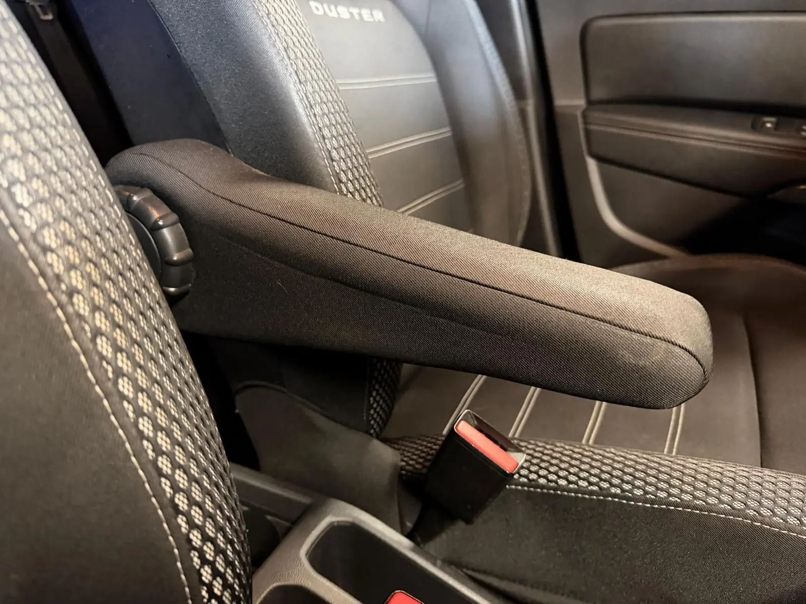 Accoudoir central relevé entre les sièges avant en tissu noir du Dacia Duster, détail intérieur.