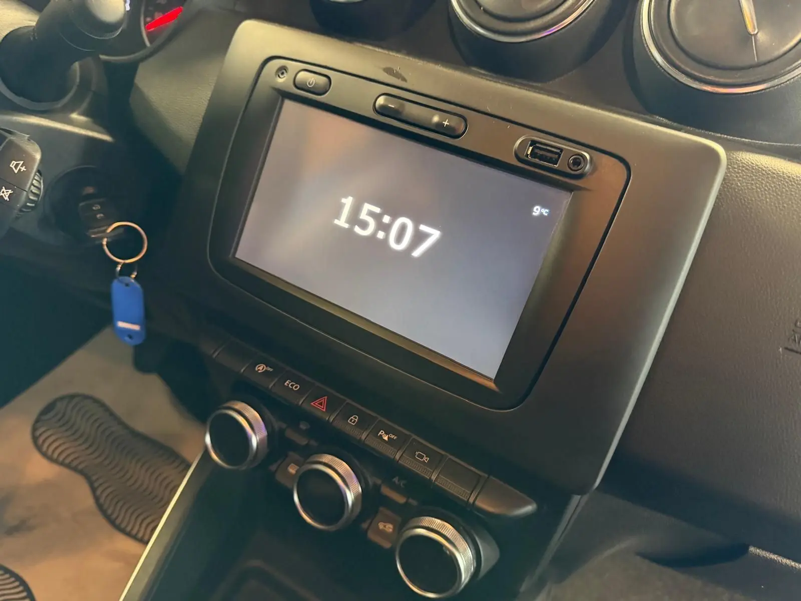 Vue rapprochée de la console centrale du Dacia Duster bleu, avec écran tactile affichant l'heure et commandes climatisation.