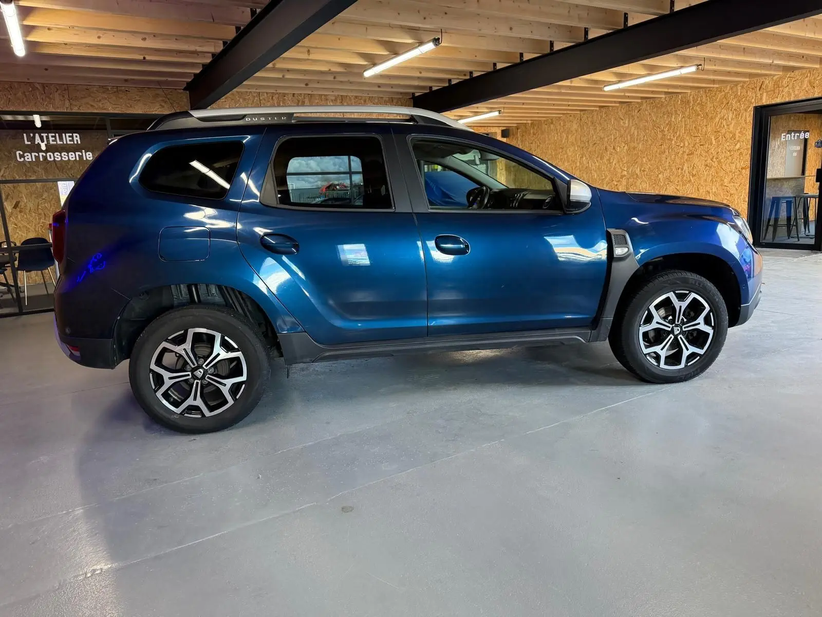 Vue latérale côté gauche d'un Dacia Duster bleu avec jantes alliage 17 pouces dans un atelier carrosserie.
