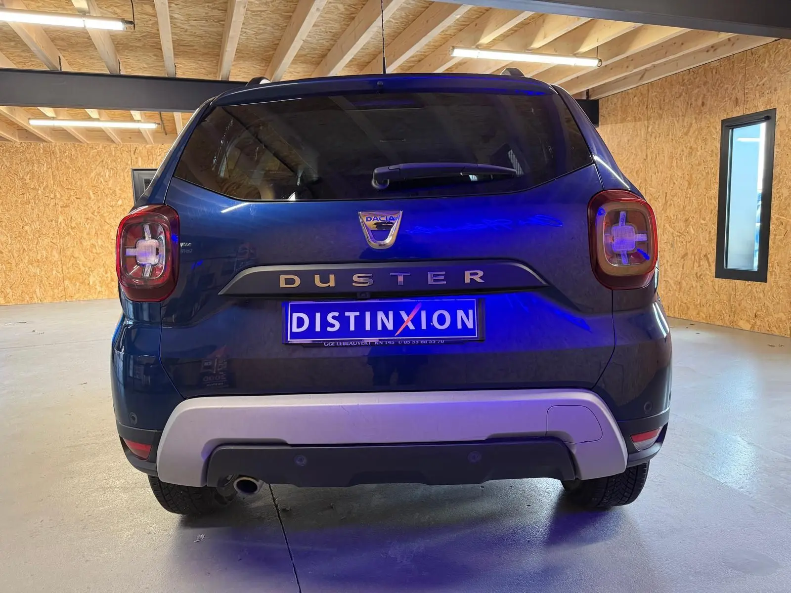 Vue arrière d'un Dacia Duster bleu avec logo distinctif et plaque d'immatriculation "DISTINXION" en intérieur lumineux.