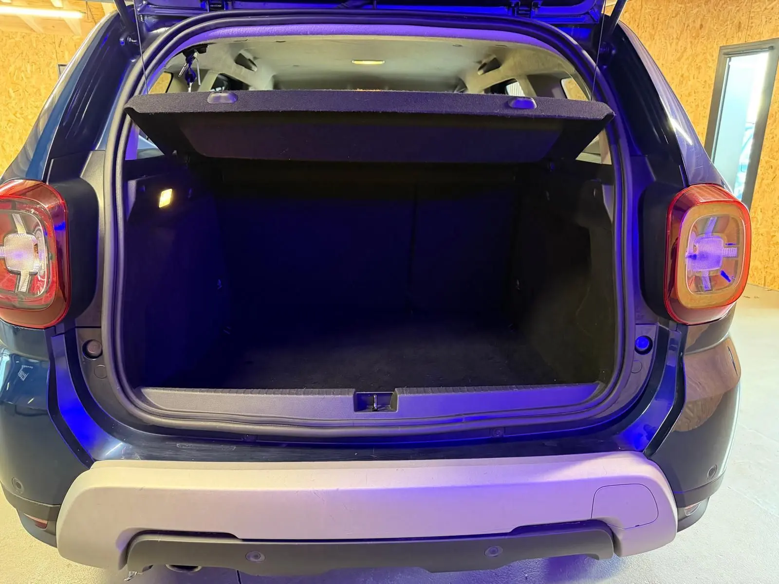 Coffre ouvert vu de l'arrière d'un Dacia Duster bleu, avec hayon et feux arrière visibles.